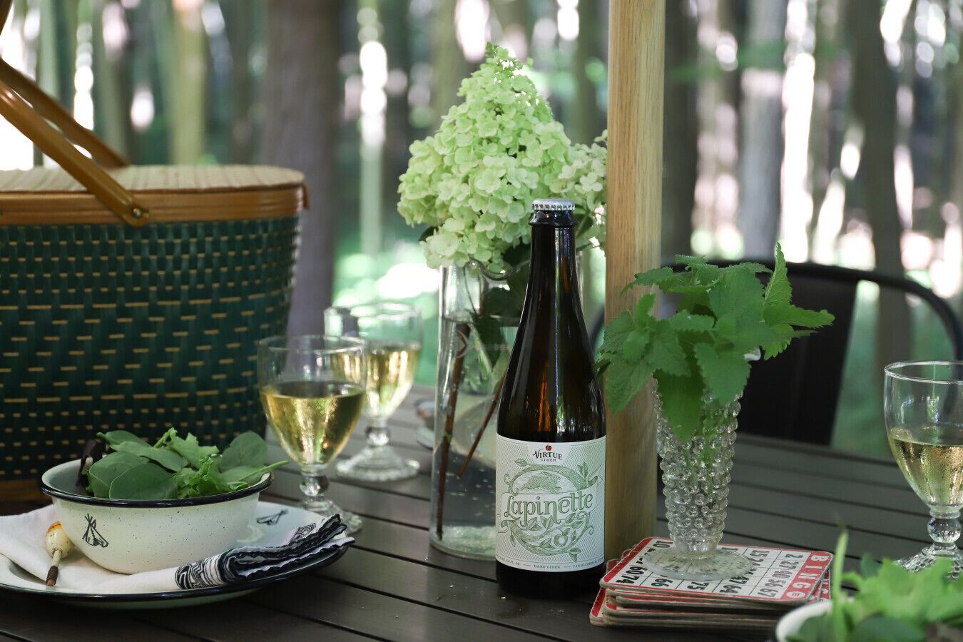 A table with a bottle of wine, glasses, a bowl of greens, and a basket.