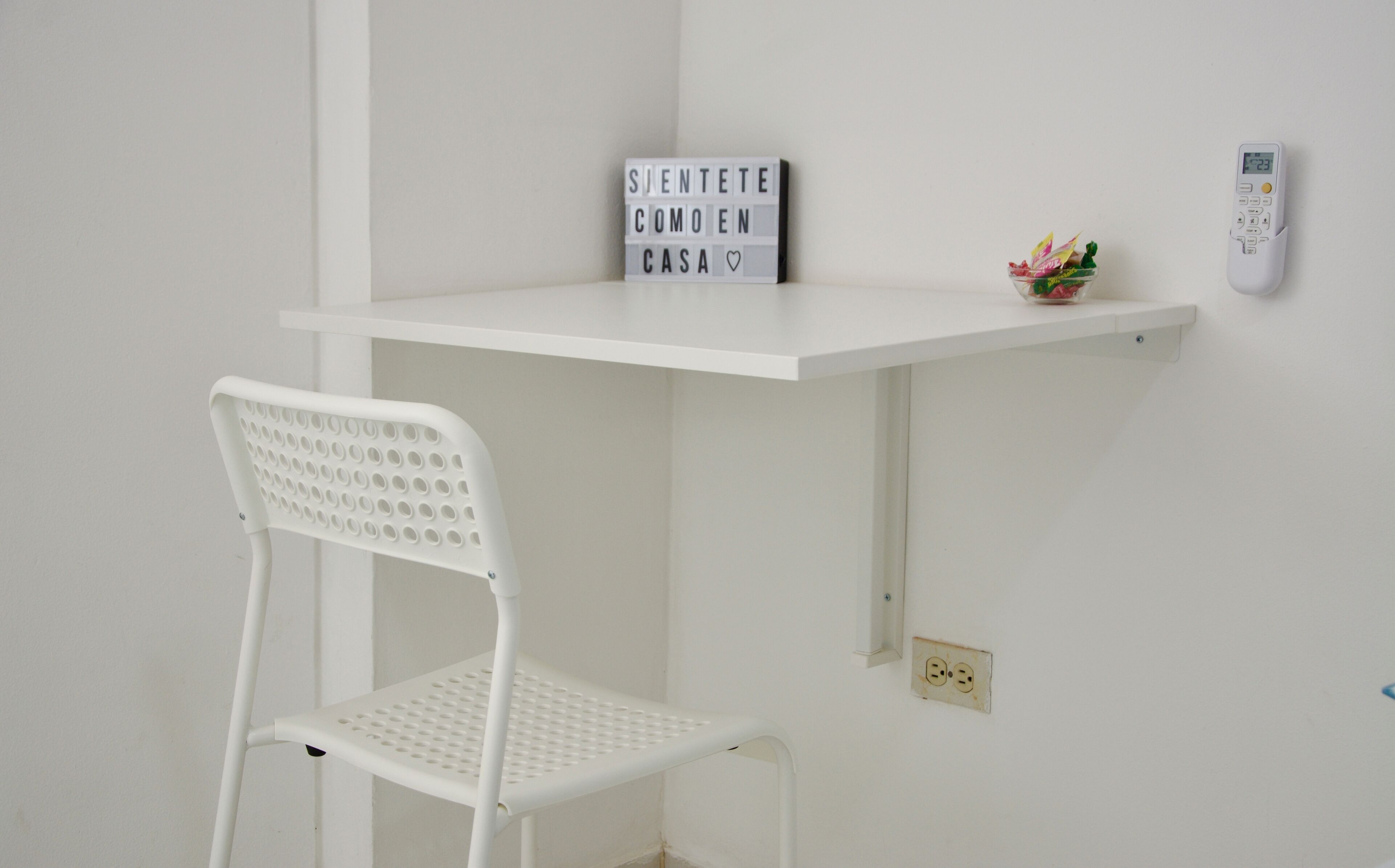 Un bureau blanc avec une chaise, une pancarte sur laquelle on peut lire « ASSEYEZ-VOUS COMME CHEZ VOUS », une petite plante et un thermostat fixé au mur.