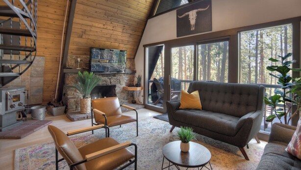 Een gezellige woonkamer met een open haard, lederen stoelen, een bank en grote ramen met uitzicht op het bos.