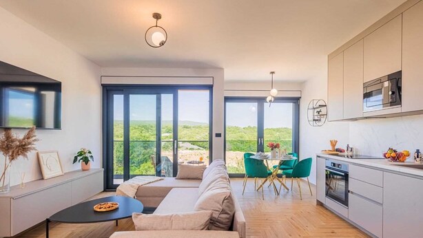 A modern living room with a sofa, a dining table, and chairs, a kitchen with a microwave and cabinets, and large windows with a view of greenery. - Luxury apartment LaRiv II with pool (Krk)