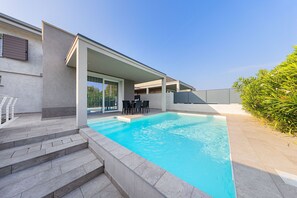 Una piscina moderna al aire libre con escalones de acceso, un área de terraza cubierta con mobiliario exterior y vistas a la vegetación circundante.