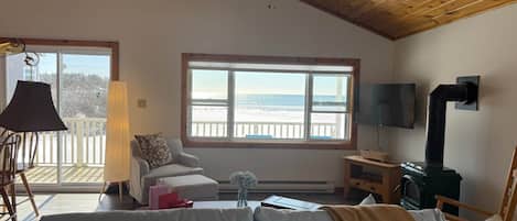 Ein gemütliches Wohnzimmer mit großem Fenster zum Strand, Deckenventilator, Kamin und einem an der Wand montierten Fernseher.