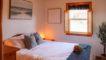 A bedroom with a bed, a bedside table, a window with shutters, a wall-mounted picture, and a potted plant.