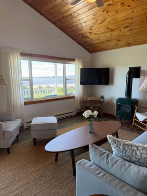 Una sala de estar acogedora con techo de madera, un ventanal con vistas, un televisor de pantalla plana, un estufa de leña verde y un cómodo mobiliario.