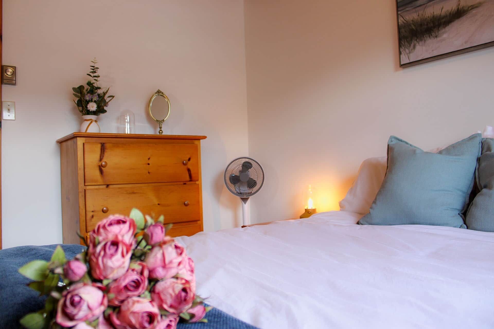 Une chambre à coucher avec une commode en bois, une table de chevet avec une lampe, un ventilateur et un bouquet de roses roses.
