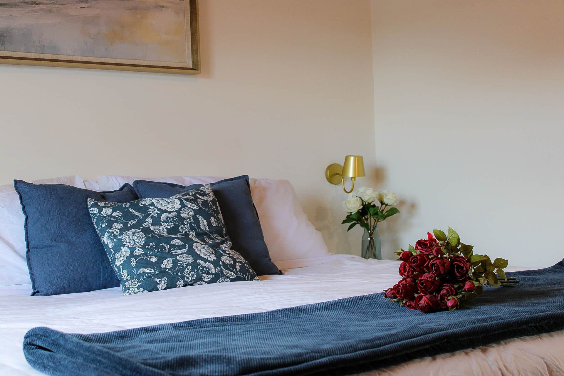 Un lit bien fait, agrémenté d’un coussin à motifs floraux, d’une couverture bleu foncé et d’un bouquet de roses rouges.