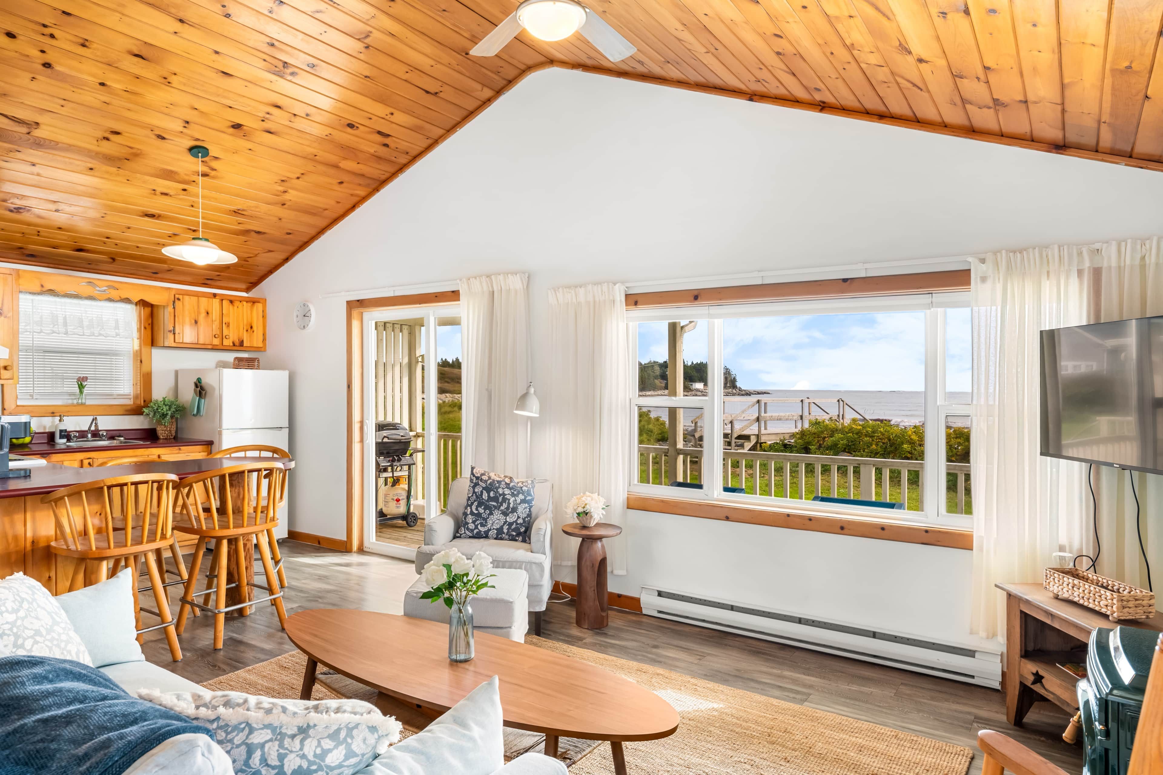 A spacious living area with a wooden ceiling, a kitchen with wooden cabinets, a dining area with chairs, a sofa, a coffee table, and a large window offering a view of the outdoors.