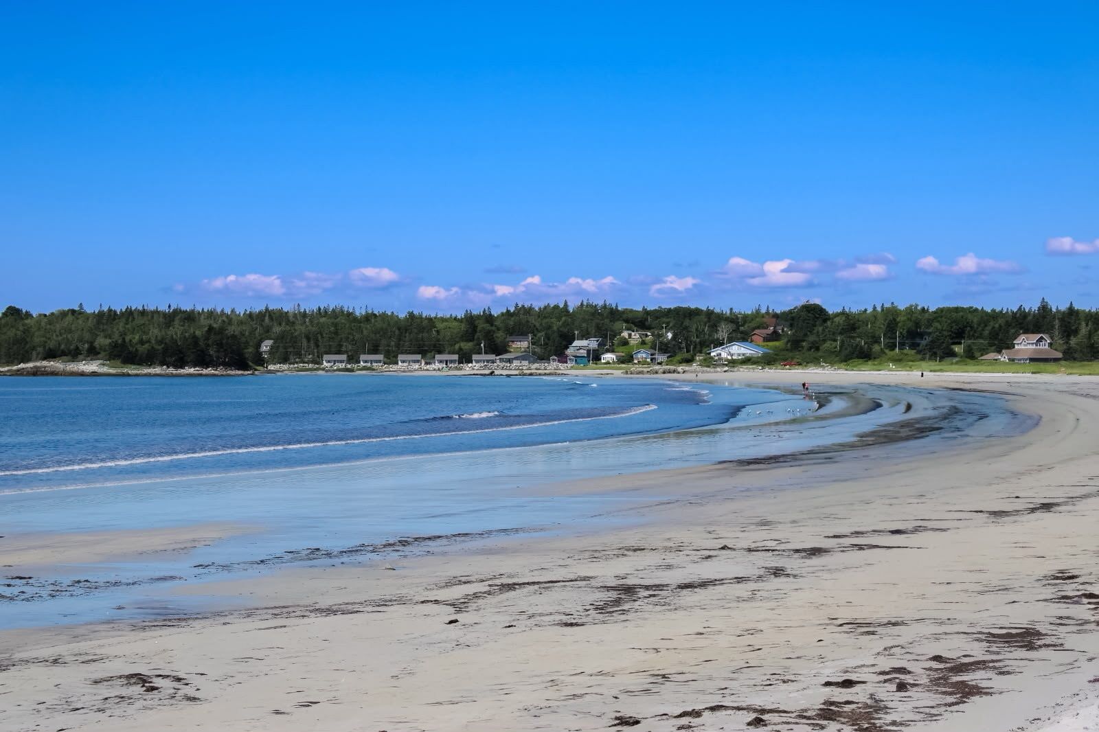 Une plage à l’eau claire, avec un rivage sablonneux et des maisons en arrière-plan.