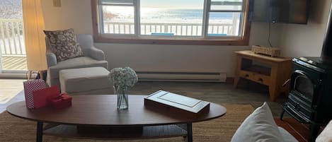 A living room with a large window overlooking a beach, a television, and a fireplace.