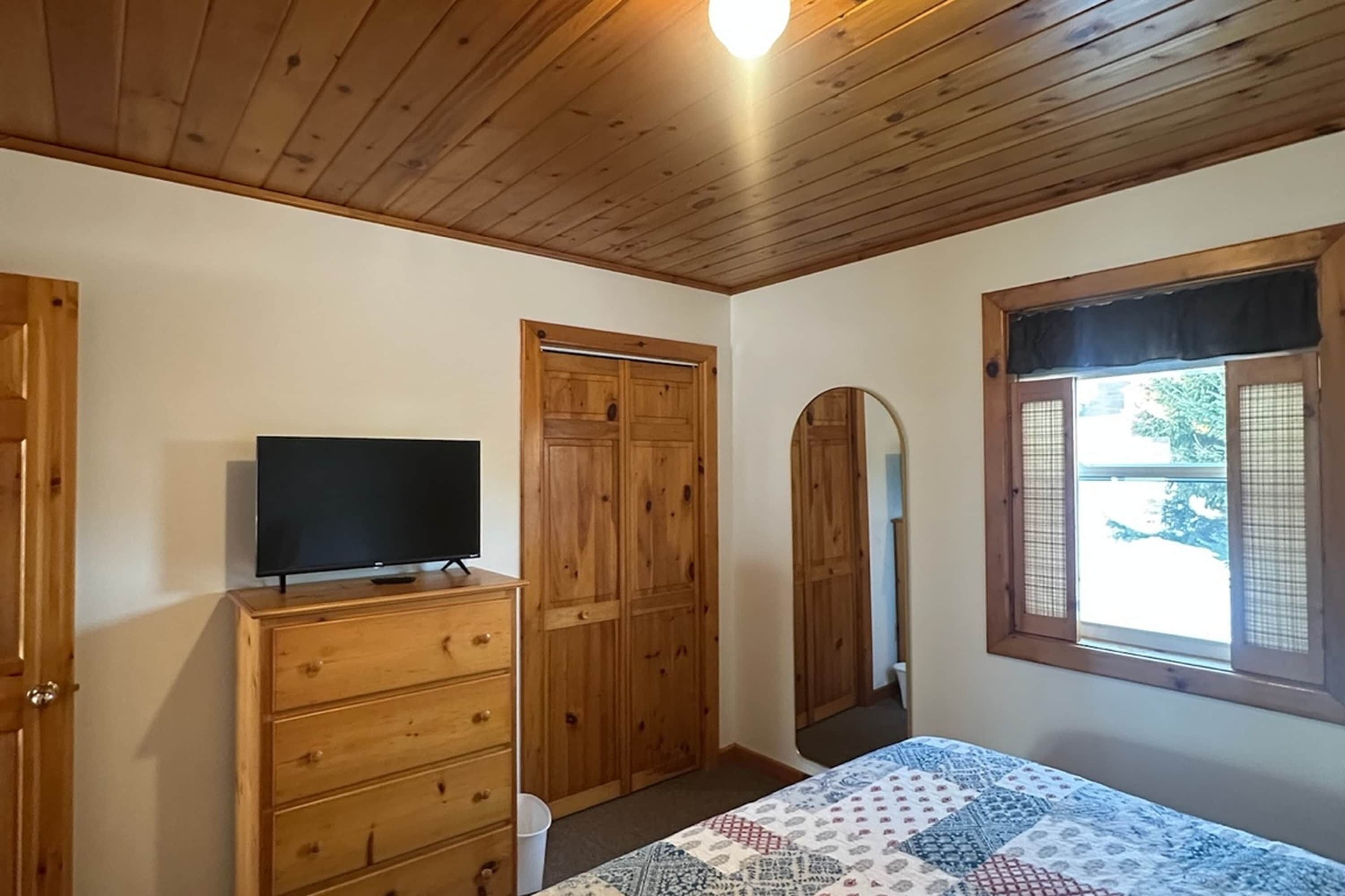 A bedroom with wooden ceiling, a bed with a patterned bedspread, a wooden dresser, a television on the dresser, and a window with a view of the outside.