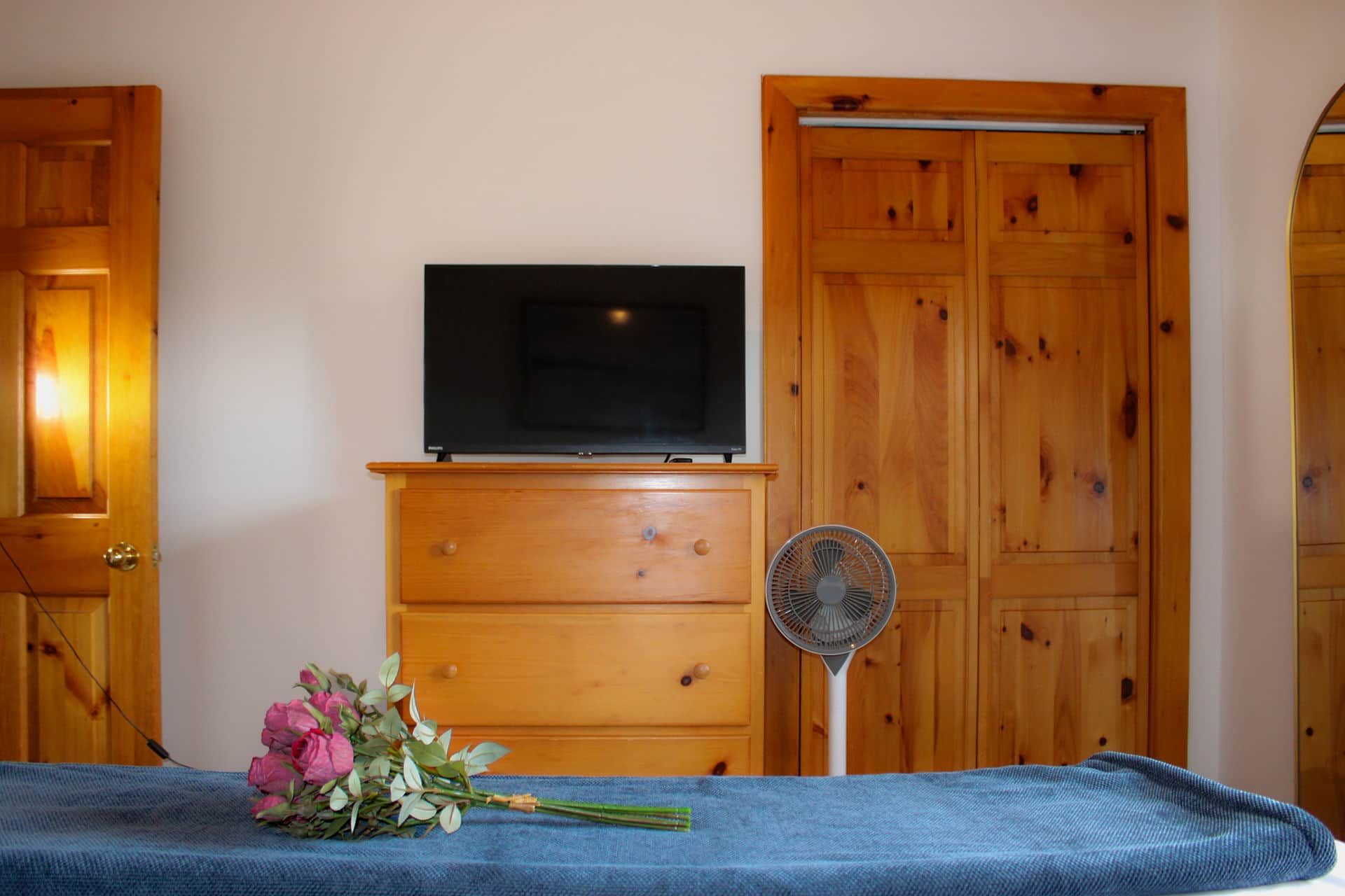 Ein Zimmer mit einem hölzernen Schminktisch, einem Flachbildfernseher darauf, einem Ventilator und einem Bett mit blauer Bettdecke.