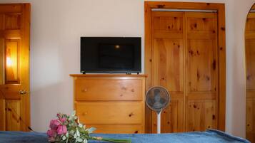 A room with a wooden dresser, a flat-screen TV on top, a fan, and a bed with a blue cover.