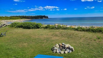 A grassy area with a fire pit and a blue mat, overlooking a calm body of water and a distant shoreline.