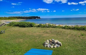 A grassy area with a fire pit and a blue mat, overlooking a calm body of water and a distant shoreline.