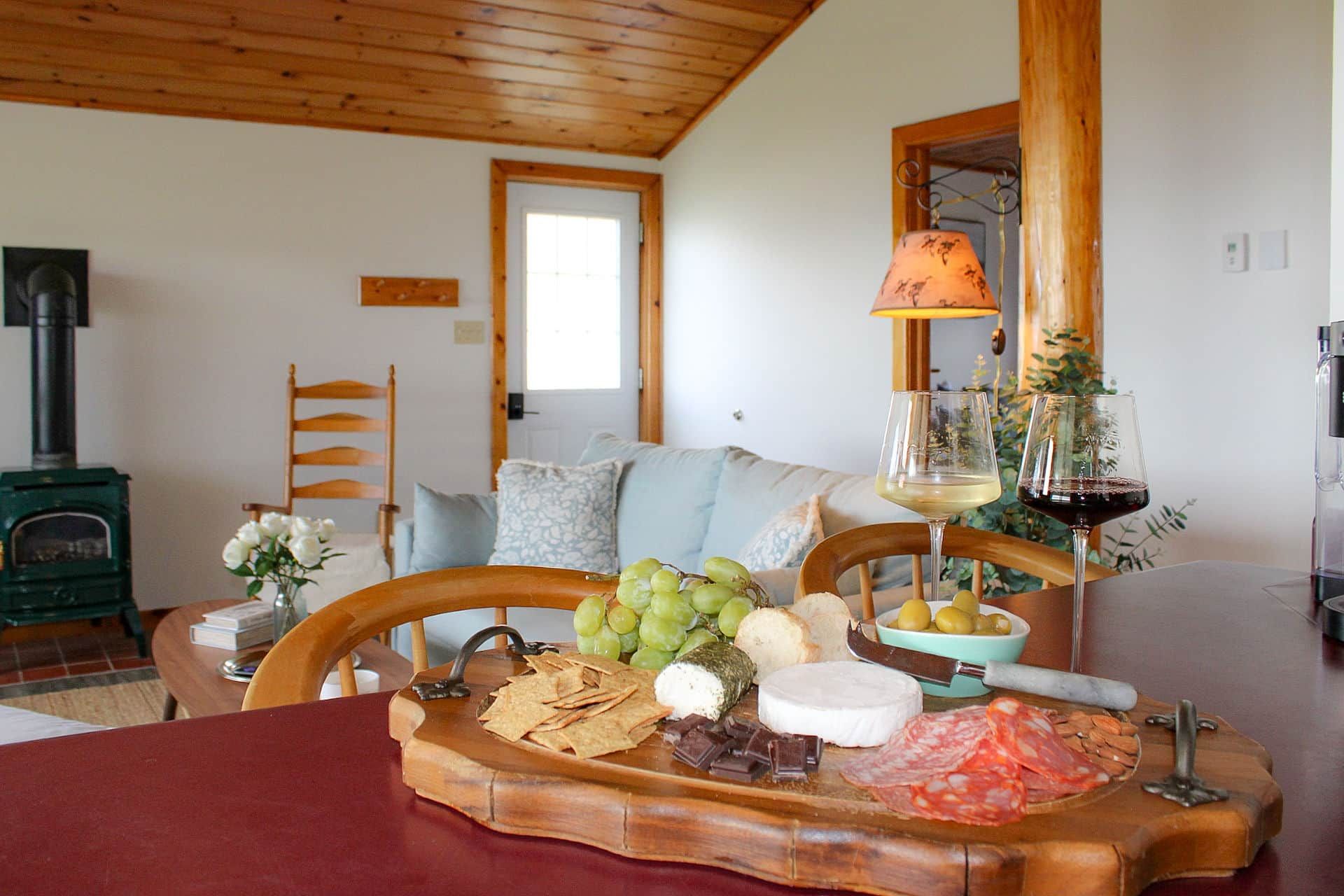 Una habitación acogedora con techo de madera, chimenea, una mesa de comedor con una tabla de quesos y una lámpara.