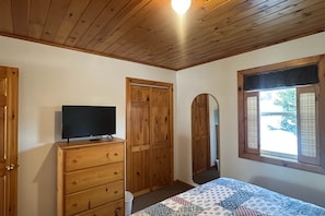 A bedroom with wooden ceiling, a bed with a patterned bedspread, a wooden dresser, a television on the dresser, and a window with a view of the outside.