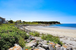 A coastal beach with a wooden walkway, houses, and a rocky shoreline.