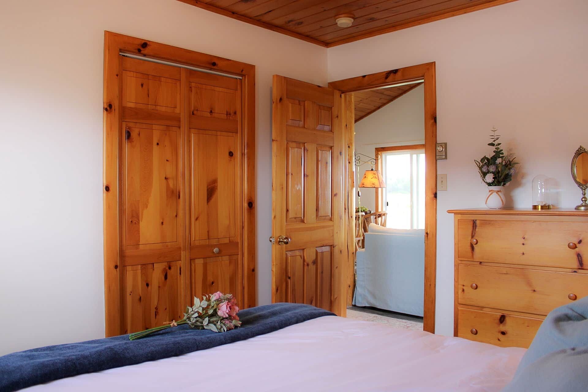 A bedroom with a wooden wardrobe, a dresser, a bed with a blue blanket, and a bouquet of flowers.