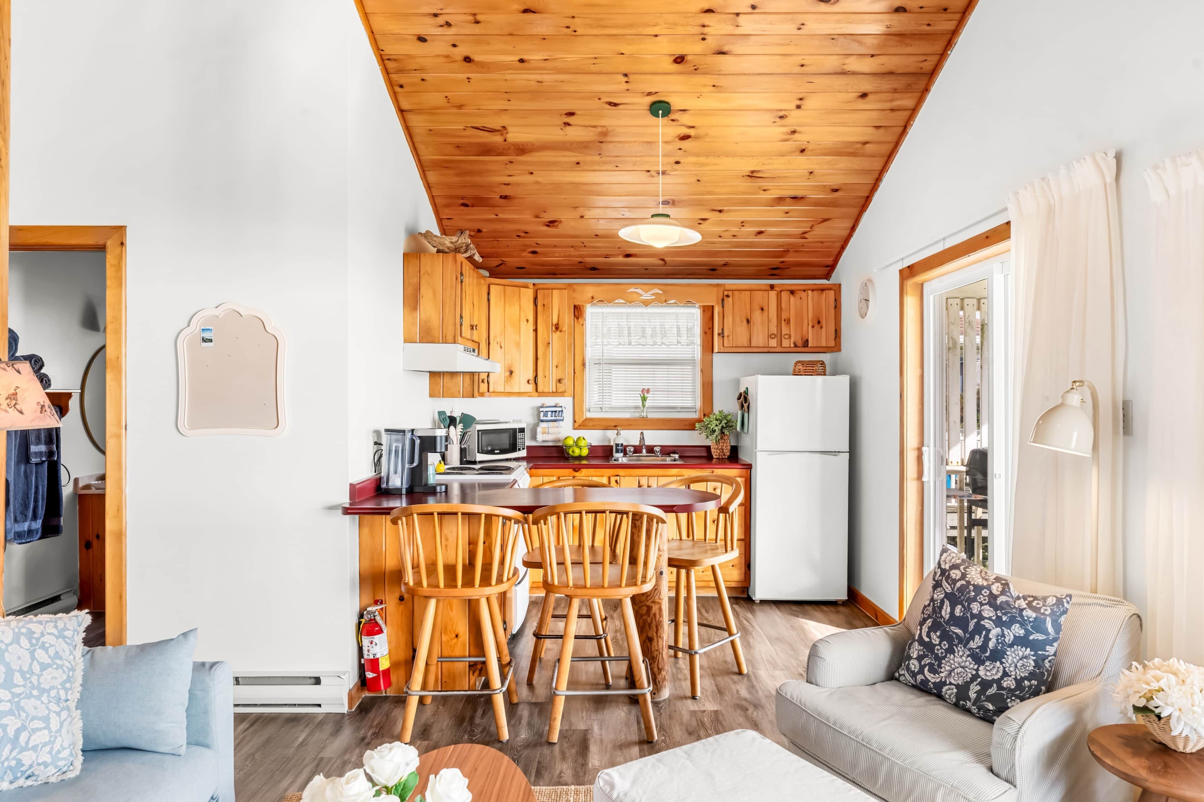 Une cuisine compacte avec des meubles en bois, un îlot central doté de tabourets, un réfrigérateur et un coin repas comprenant un canapé et des chaises.