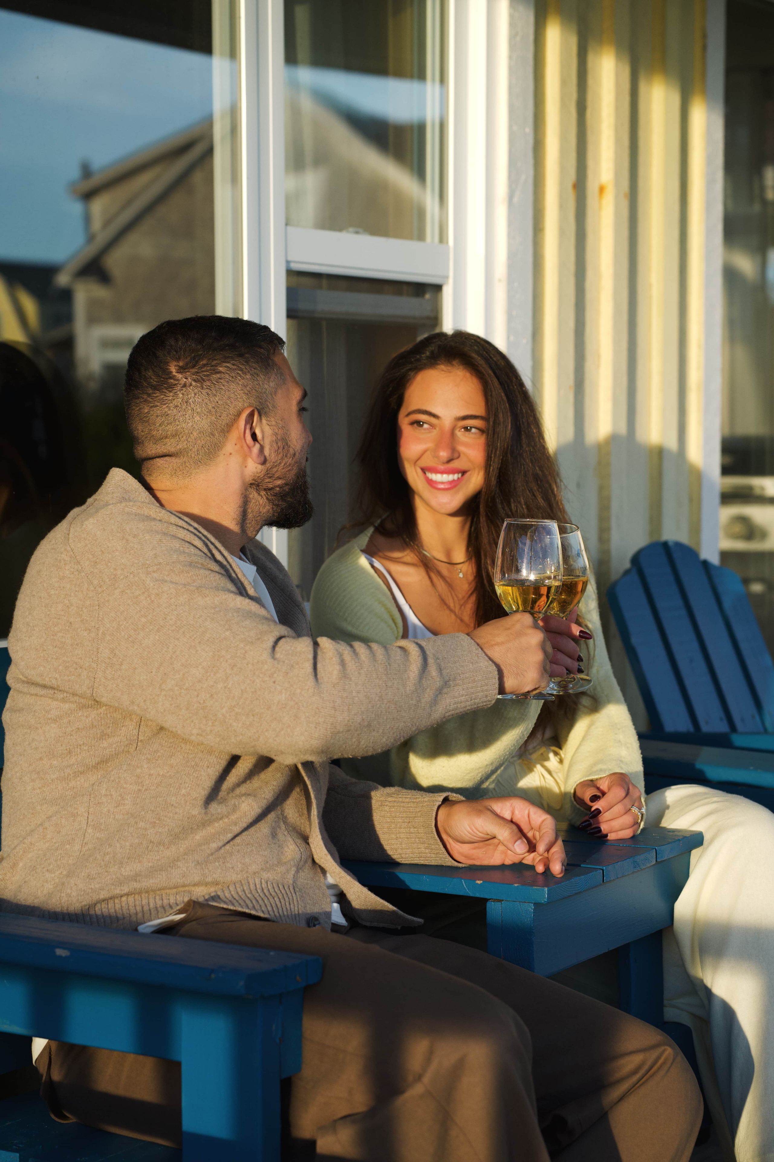 A couple seated at an outdoor table with a glass of wine, a blue chair, and a window in the background.