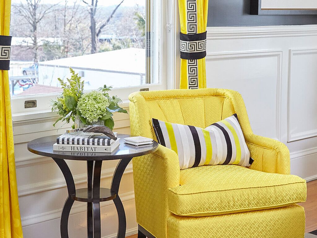 A yellow armchair with a striped pillow, a small round table with a book and a plant, and a window with a view of trees and a building.