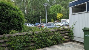Een parkeerplaats met auto's, een houten hek en een groene afvalcontainer.