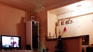 Un salon avec une télévision, une table à manger avec un plateau, un sapin de Noël et une cuisine en arrière-plan.