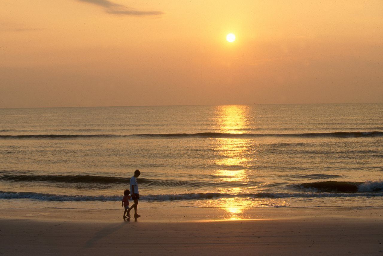 A sunset over a beach with gentle waves and a person walking on the sand.