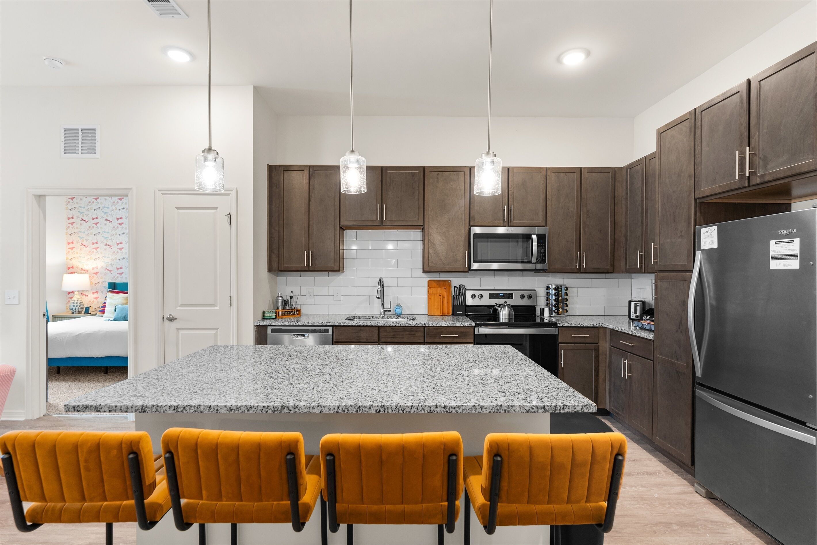 A modern kitchen with granite countertops, dark wooden cabinets, a stainless steel refrigerator, and a central island with four yellow chairs.