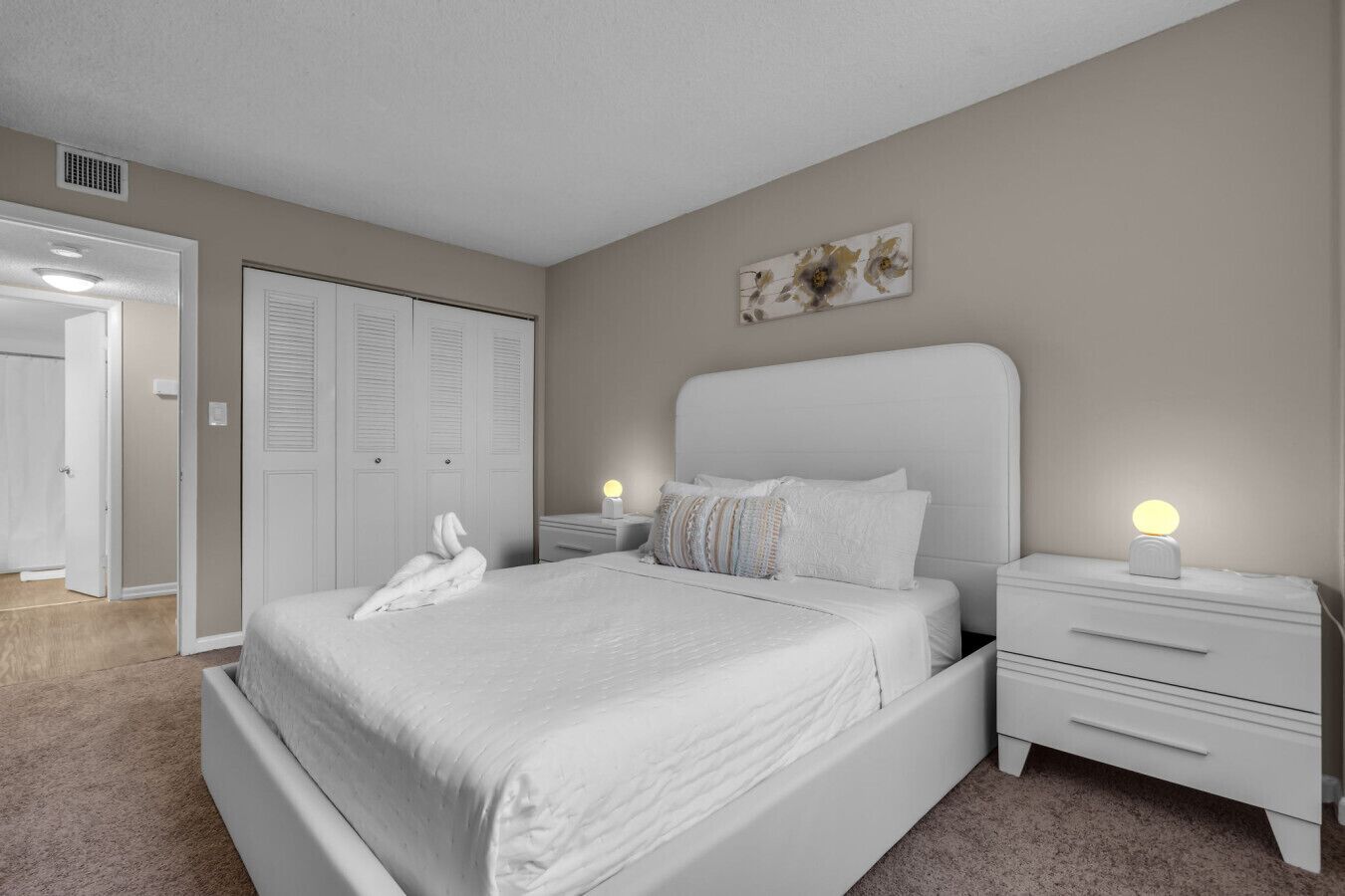 Un dormitorio con una cama grande, dos mesitas de noche con lámparas, un armario y una puerta que da a otra habitación.