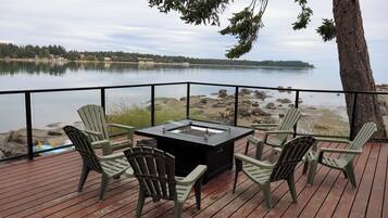 A deck with a fire pit table and chairs, overlooking a lake with a rocky shoreline.