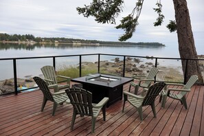 A deck with a fire pit table and chairs, overlooking a lake with a rocky shoreline.