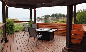Eine Holzterrasse mit Tisch und Stühlen, mit Blick auf ein Gewässer und eine entfernte Küstenlinie.