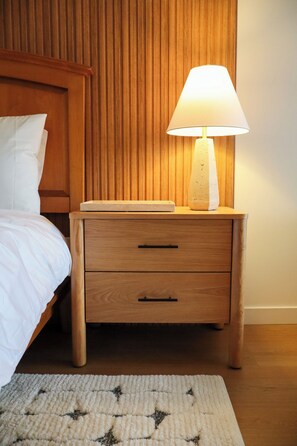 A wooden bedside table with a lamp, a bed with white bedding, and a patterned rug on the floor.