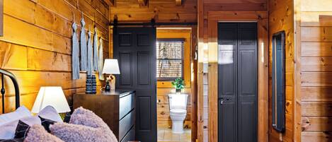Interior de una cabaña de madera con una cama, un aparador, una lámpara y vistas al baño por una puerta abierta.
