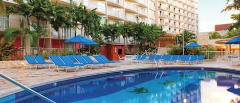 Un espace piscine d’hôtel avec des chaises longues bleues et des parasols, entouré de palmiers et d’un immeuble à plusieurs étages.