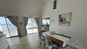 Un comedor con una mesa de madera y sillas blancas, ventanas amplias con cortinas, y vista a la playa.