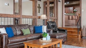 A spacious living room with a brown leather sofa, a wooden coffee table, and a black leather chair. A wooden staircase leads to a kitchen area.