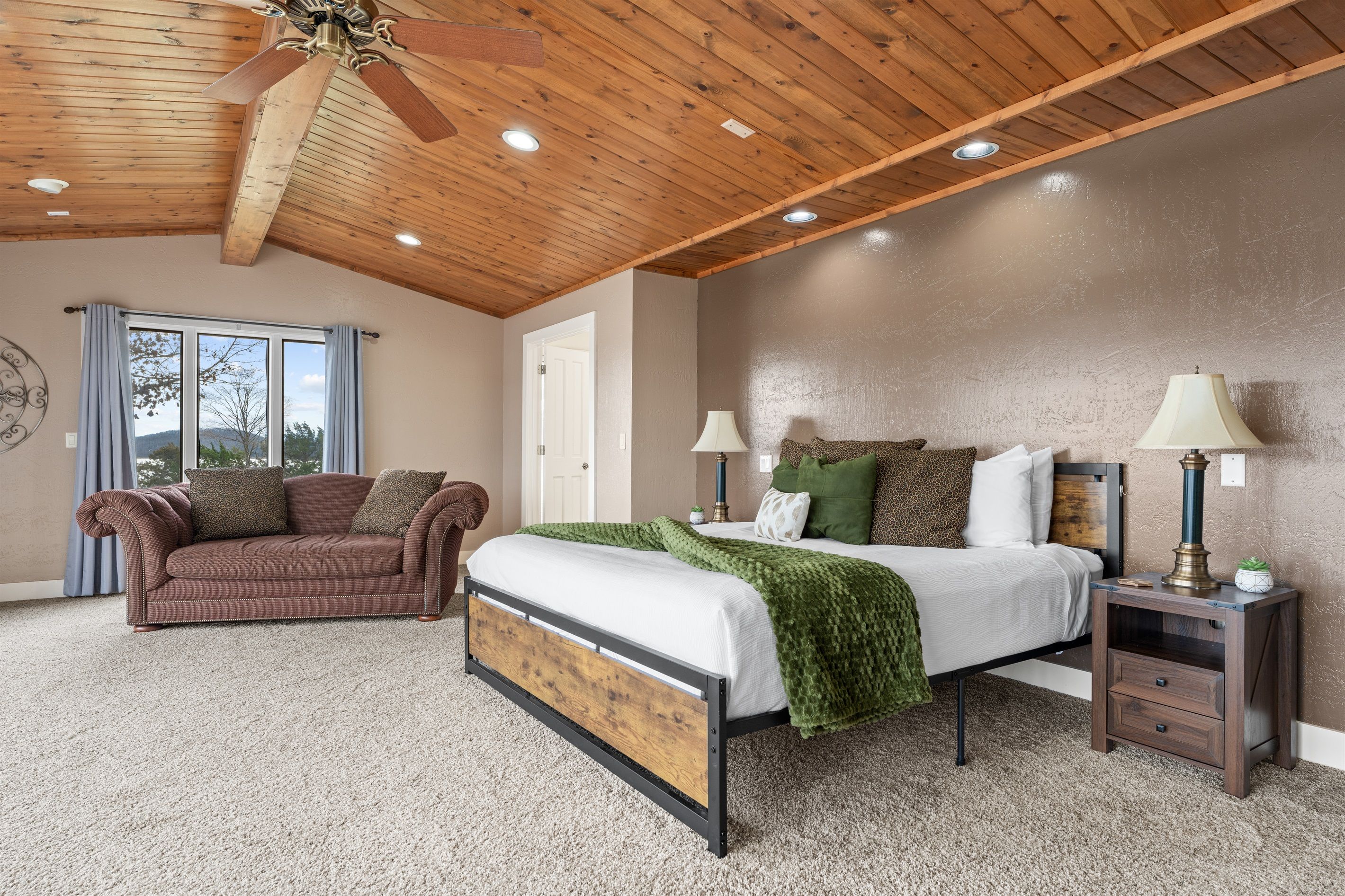 A bedroom with a wooden ceiling, a bed with a green blanket, a nightstand with lamps, and a window with curtains.