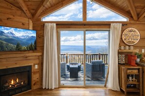 Un intérieur de chalet chaleureux avec une cheminée, une grande porte-fenêtre coulissante donnant sur une terrasse extérieure avec un foyer et des sièges, et une vue panoramique sur les montagnes.