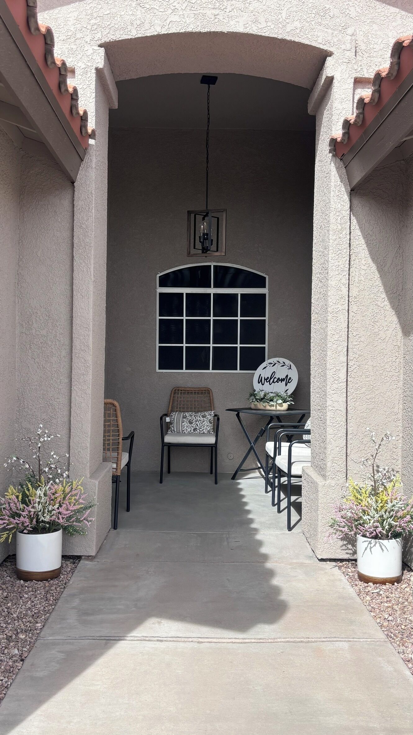 Un patio cubierto con un letrero de bienvenida, dos macetas, una silla de mimbre y una mesita.