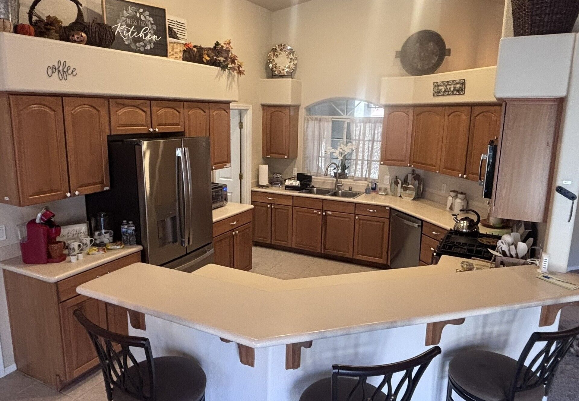 Una cocina con armarios de madera, un refrigerador de acero inoxidable, una isla central con sillas y una ventana con cortinas.