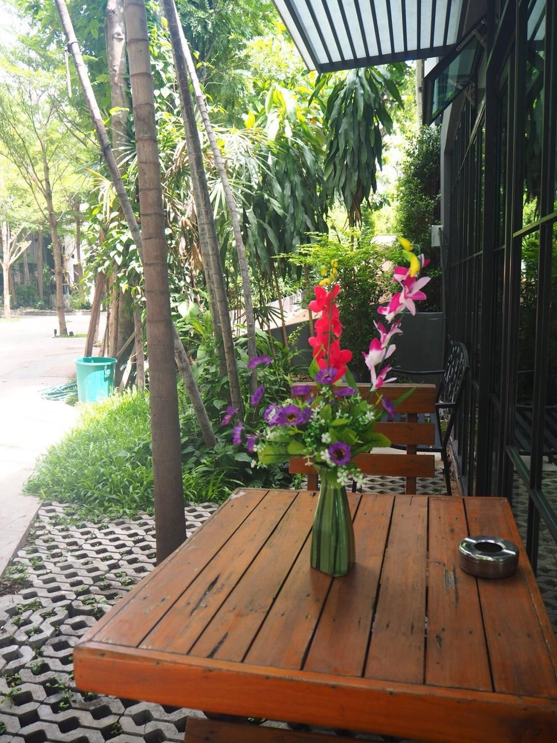 An outdoor wooden table with a vase of flowers, surrounded by greenery and a metal fence.