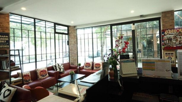 A cozy lobby with a red leather sofa, a coffee table, and a bookshelf. Large windows provide natural light and a view of greenery outside. - BB House Chaingmai (Chiang Mai)