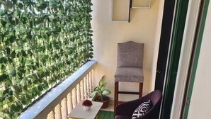 Uma varanda com piso de grama artificial verde, uma mesinha com um vaso de planta, duas cadeiras e uma janela com vista para o verde.