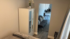 Ein kleiner Wohnbereich mit einem Bett, einem kleinen Schrank und Blick in einen Küchenbereich mit einer Couch und Barhocker.