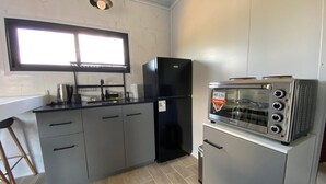 Cocina compacta con refrigerador negro, horno tostador empotrado, ventana sobre el lavamanos y un taburete de madera.