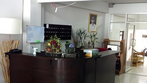 A reception desk with a dark wood finish, a whiteboard with text, a plant arrangement, and a framed picture on the wall. - S K Airport (Chiang Mai)