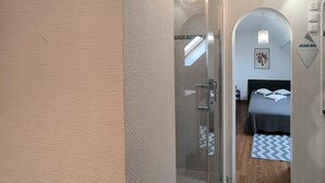 Una habitación de hotel moderna con una puerta de cristal, una cama con colcha gris, suelo de madera y un candelabro.