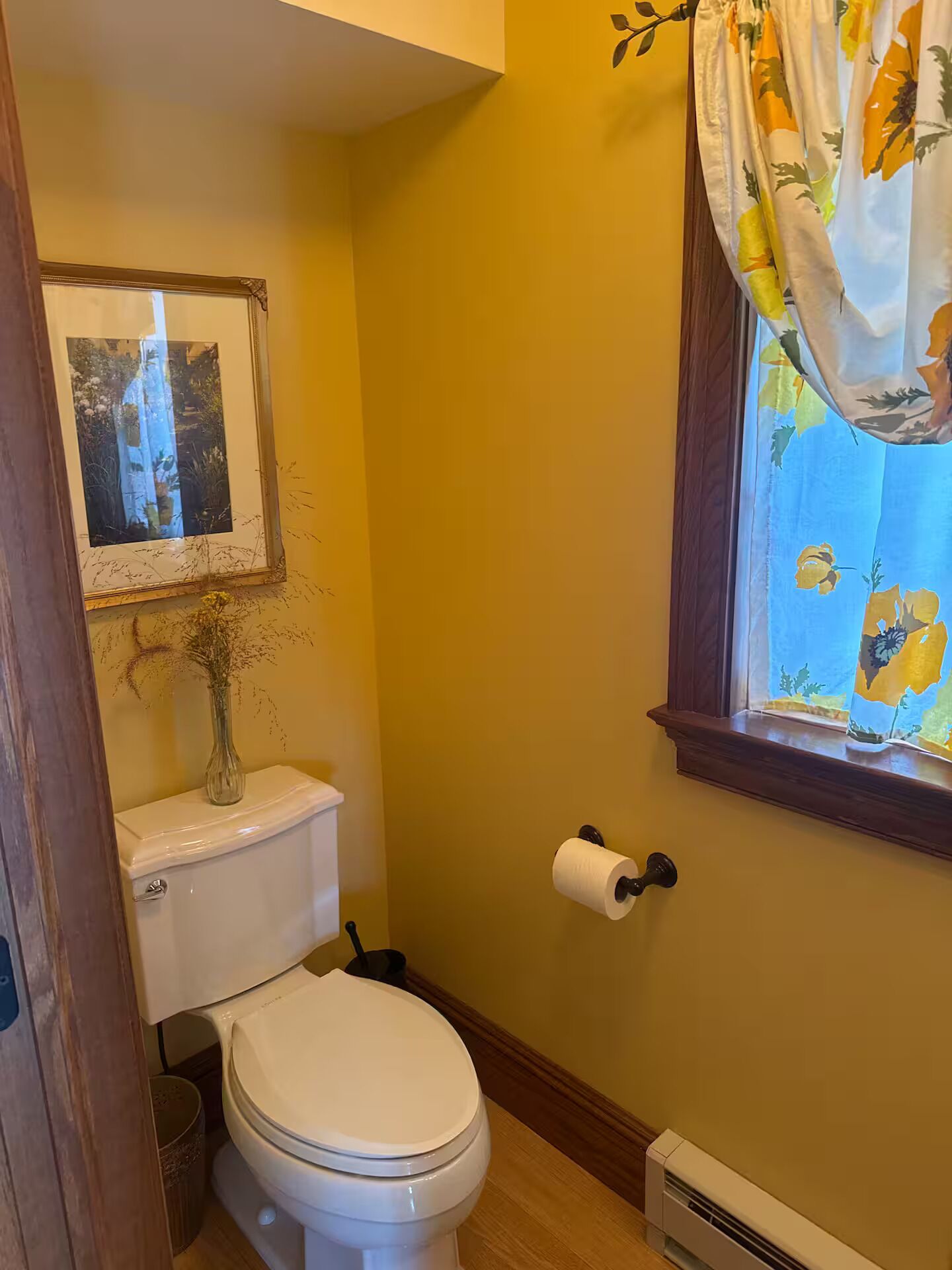A bathroom with a toilet, a picture frame, a vase, a window with floral curtains, and a wall-mounted toilet paper holder.